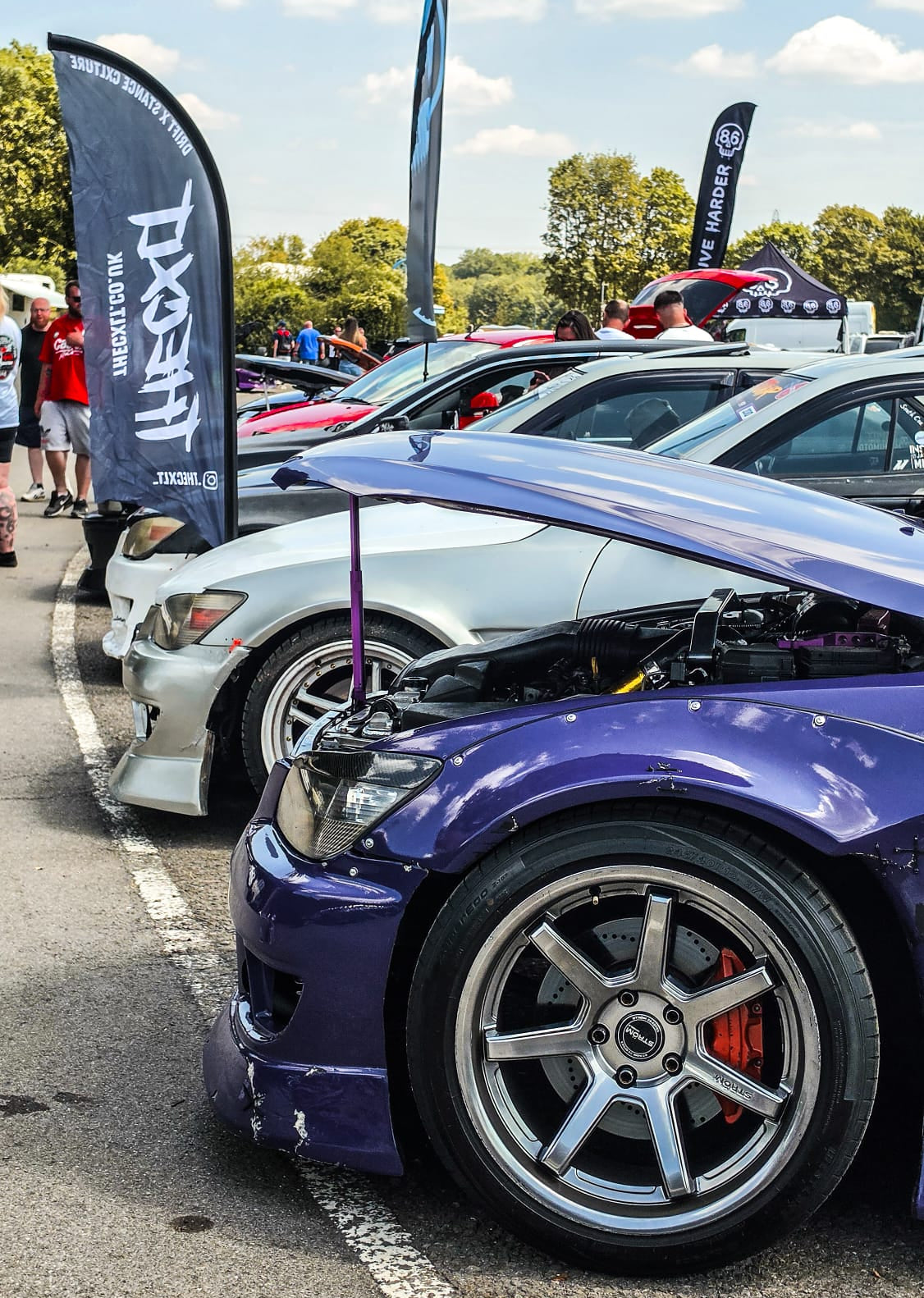 A close-up outdoor shot of a purple BMW sedan with its hood open, parked at an outdoor car show. Other cars, event flags, and a crowd are visible in the background under a blue sky.